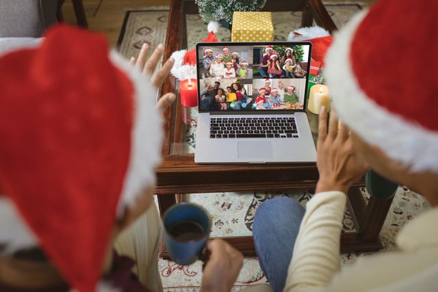 Diverse couple wearing santa hats enjoying a virtual video call with friends during Christmas celebration. Scene includes festive decorations, laptop showing various smiling friends, and candles. Ideal for use in articles or campaigns about digital holiday celebrations, virtual gatherings, and modern communication during festive seasons.