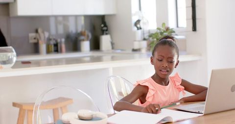African girl studying at kitchen table with laptop and notebook, concentrating on homework
