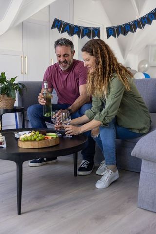 Mature couple celebrating birthday at home pouring wine and sharing cheese platter on sofa