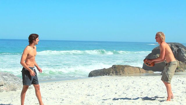 Two friends playing rugby on a sunny beach with an ocean view. Ideal for use in travel and tourism publications, sports and activity promotions, summer camp advertisements, or social media content focused on outdoor activities and fitness.