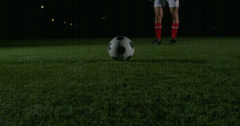 Hovering soccer ball over wet pitch at night while player standing in red knee-high socks