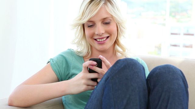 Young blonde woman wearing casual clothes sitting comfortably while texting on her smartphone. Ideal for themes related to modern lifestyle, communication, indoor activities, and leisure time. Suitable for use in advertising, editorial stories, website banners, and social media content aiming to represent youthful relaxation and connectivity.