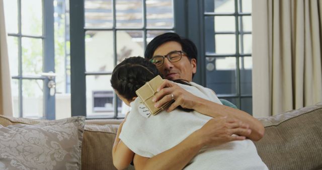 A middle-aged Asian man is embracing a young girl, his daughter, in a warm hug in a cozy living room setting, with copy space. They share a moment of affection, enhancing the sense of family bonding and love.