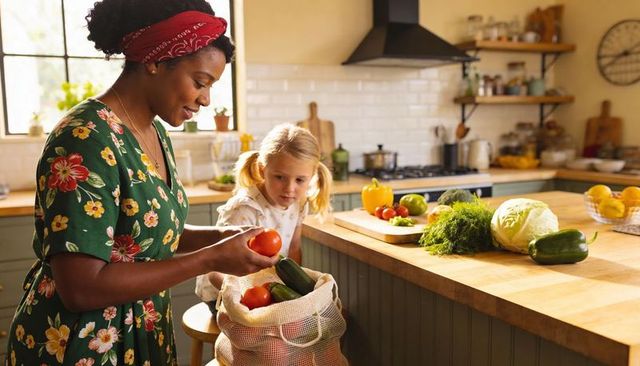 Woman unpacking fresh vegetables into reusable mesh bag at sunlit kitchen while child watches