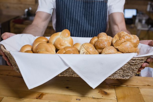 Waiter holding a basket filled with freshly baked pastries, including bread rolls and sesame seed buns, at a wooden counter in a cafe. Ideal for use in content related to cafes, bakeries, hospitality, food service, and fresh baked goods.