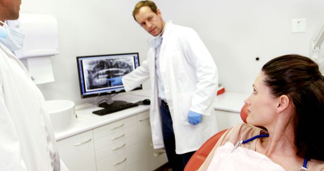 Dentist in white coat points at dental X-ray on computer screen while explaining condition to female patient sitting in chair. Concept of professional dental care, patient-doctor communication, and use of technology in dentistry. Suitable for imagery promoting dental health services, clinics, or educational materials related to dentistry and patient care.