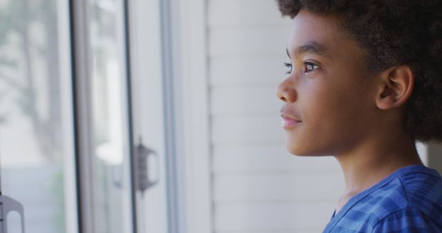 A young African American boy is looking out a window, appearing thoughtful, dressed in a blue shirt. He stands by the window in natural daylight, creating a calm and reflective mood. This can be used for concepts such as childhood dreams, contemplation, indoor activities, innocence, and home life.