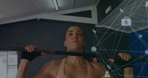 Female athlete pulling up on bar in industrial gym wearing pink top and wrist wraps
