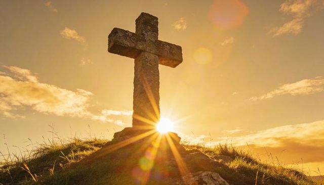 Weathered stone cross glowing with sunburst on grassy hilltop at golden hour