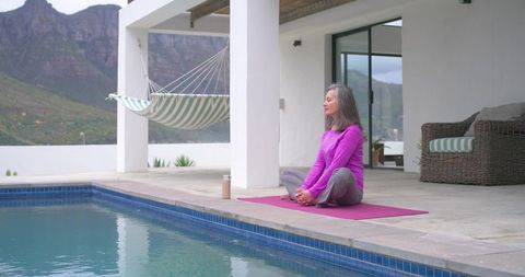 Mature woman practicing poolside yoga meditation on patio mat with hammock and mountain view