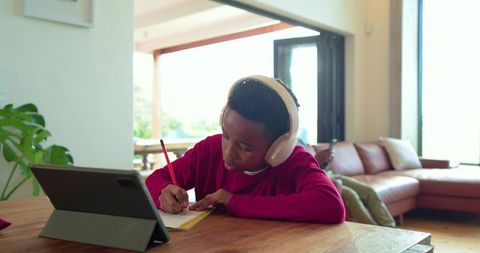 African american boy studying at home with tablet and headphones, writing notes in sunlit lounge