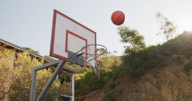 This image shows a basketball in mid-air approaching a hoop with the backboard and net clearly visible. The scene is set outdoors against a natural backdrop that includes trees and hillside. Could be used for sports-related projects, illustrating athletic activity, promotional material for youth sports leagues or outdoor game advertisements.