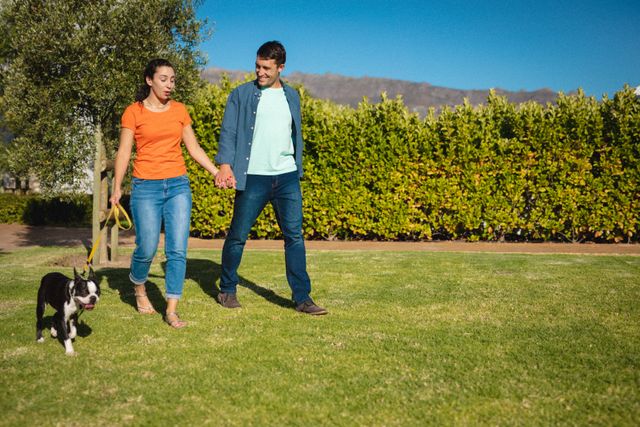 Caucasian couple enjoying a sunny day walking their dog in a lush garden. Ideal for use in advertisements promoting pet care products, outdoor activities, or lifestyle blogs focusing on relationships and leisure.