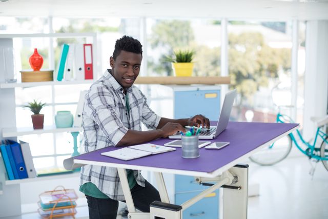 Young professional working on laptop in modern office environment. He is smiling and appears productive. The setting includes various office furnishings such as bookshelves, binders, and plants. Suitable for use in articles about modern office environments, productivity, workplace diversity, and business technology.