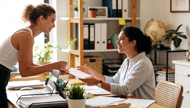 Colleagues exchanging documents while smiling at bright modern shared workspace teamwork