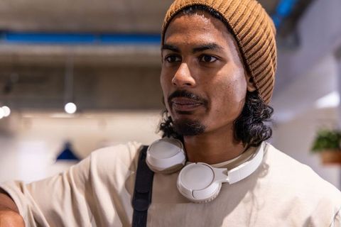 Asian man in 20s leaning in coworking space wearing brown beanie and white headphones