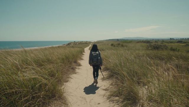 Woman walking coastal dune path with backpack and tripod, minimalist seaside hiking scene