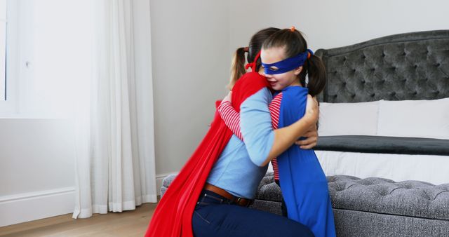 Happy caucasian mother and daughter in superhero masks and capes hugging in bedroom, copy space. Love, dressing up, childhood, motherhood, togetherness, free time and domestic life, unaltered.
