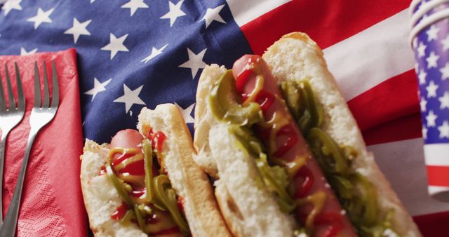 Two hot dogs with mustard and ketchup placed on top of an American flag. Red napkin and silverware add to the festive presentation with a Stars and Stripes cup in the background suggesting a patriotic celebration. Perfect for illustrating American holidays like Independence Day, BBQ invitations, summer events, or patriotic themed parties.