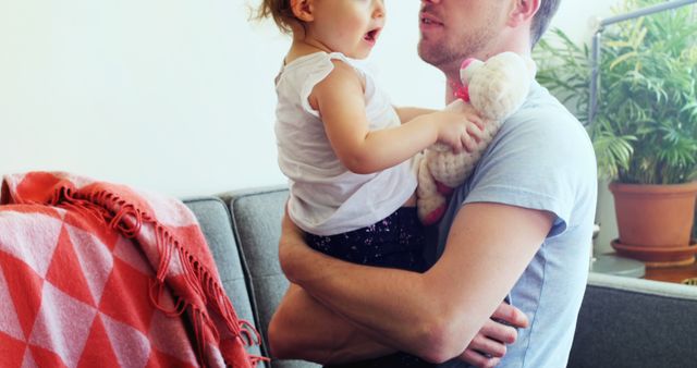 A young Caucasian father holds his toddler daughter in a cozy home setting, with copy space. Their bond is evident as she clutches a stuffed toy while enjoying a loving embrace.
