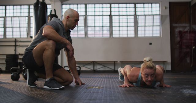 Personal trainer kneels on gym mat guiding senior woman who performs push-ups. Bright, spacious gym with fitness equipment in background. Ideal for use in articles and advertisements on fitness training, senior health, personal coaching, active lifestyle promotion, and strength training exercises for older adults.