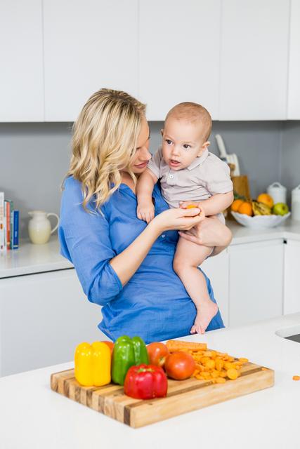 Mother holding her baby while standing in a modern kitchen, surrounded by fresh colorful vegetables. Ideal for use in family blogs, parenting articles, advertisements for home appliances, and healthy eating promotions.