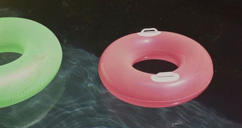 Sunlit pink and green inflatable rings floating on dark pool water with reflective ripples