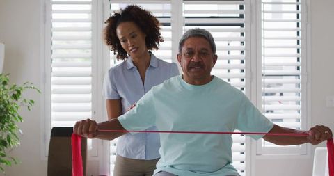 Senior Man Doing Resistance Band Shoulder Exercise with Caregiver in Home Rehabilitation