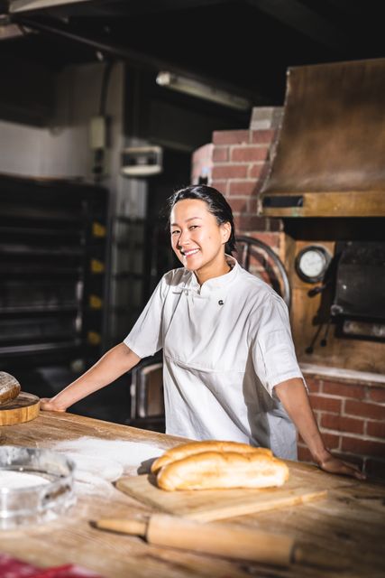 Portrait of smiling hispanic female baker standing at counter with dough and four in kitchen. unaltered, blue-collar worker, skilled, food and drink industry concept.