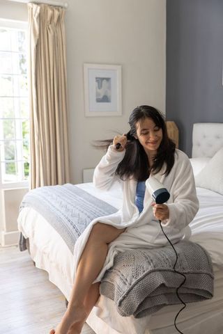 Asian woman styling hair with hair dryer in cozy bedroom wearing white bathrobe