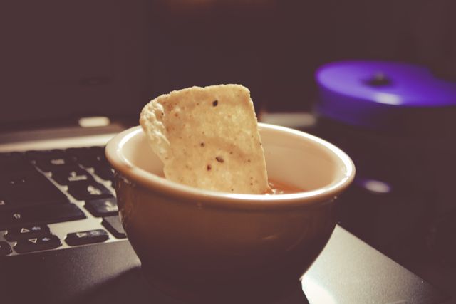 This close-up shows a tortilla chip dipped in salsa, placed in a decorative bowl on a laptop. Ideal for illustrating remote work culture, snack time during work, or promoting Mexican food appetizers. Perfect for websites, advertisements, and blogs about food, work, or casual relaxation moments.