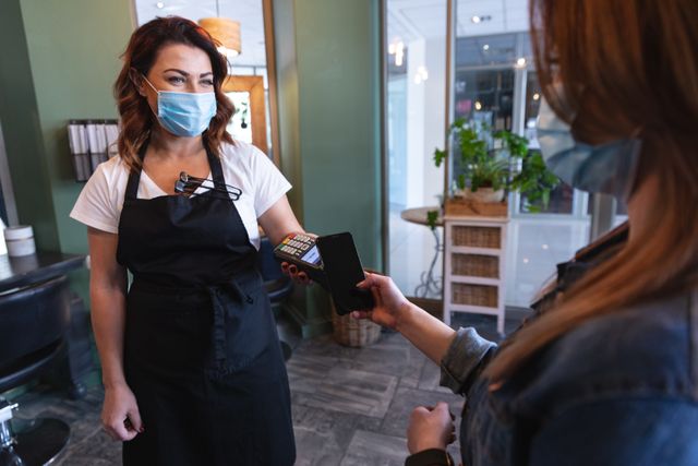 Caucasian female hairdresser in hair salon, customer making contactless payment with smartphone, wearing face masks. Health and hygiene in workplace during Coronavirus Covid 19 pandemic.