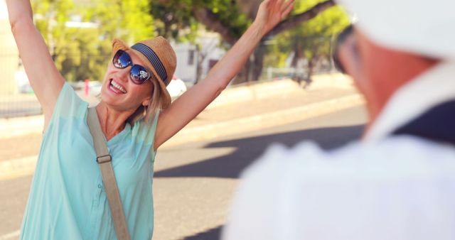 Woman enjoying vacation, smiling broadly with raised arms, wearing summer clothes, hat, and sunglasses. Captured on a sunny day in an urban outdoor setting. Ideal for travel blogs, vacation advertisements, and lifestyle promotions highlighting joy and leisure.