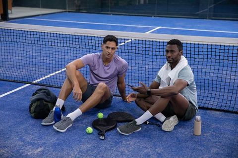 Two male players resting on blue pickleball court by net with paddles, balls and water bottles