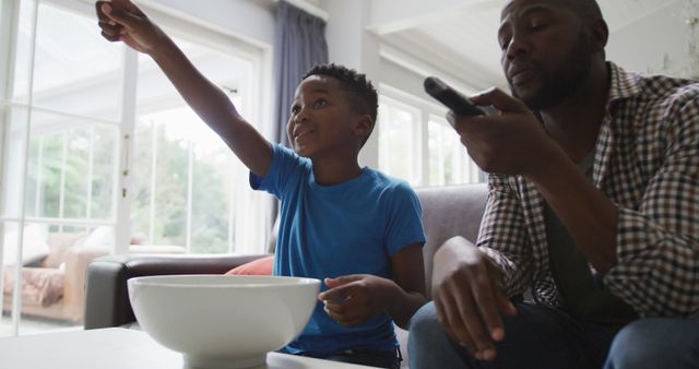 Father and son spending quality time together in a bright living room environment. Father holding a remote control while son enthusiastically gesturing and enjoying a snack. Great for concepts like fatherhood, family bonding, relaxation, and leisure activities.