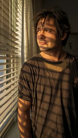 Vertical video of man tilting head into golden light through blinds casting bold shadow stripes