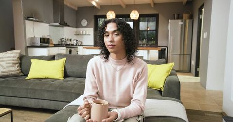 Non-binary person relaxing on gray sofa holding mug in modern minimalist kitchen