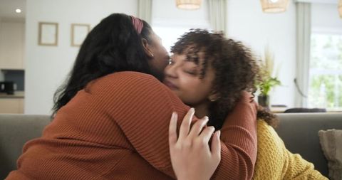 Friends hugging on sofa in sunlit living room wearing knit sweaters showing warm affection and manic