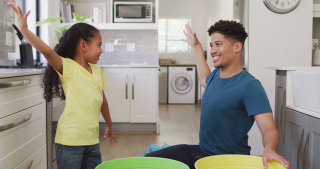 Happy father and daughter enjoying playful moments together in modern kitchen. She is wearing a yellow t-shirt and black curly hair, he is in a blue t-shirt. Great depiction of family bonding, joy, and positive domestic living. Ideal for use in family-oriented marketing campaigns, articles on parenting, and lifestyle blogs.