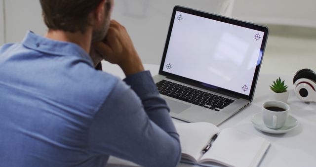 Man wearing blue shirt sitting at desk and working on laptop. Desk includes coffee cup, notebook, and headphones. Suitable for topics on remote work, productivity, modern office environment, and professional lifestyles.