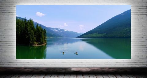 canvas print displaying two kayakers on a serene lake surrounded by mountains and trees, creating a natural ambiance. the canvas is set against a white brick wall, ideal for showcasing home or office decor potential. useful for illustrating both adventurous travel and interior design inspiration, suitable for usage in advertisements, blogs, and catalogs related to home garden decor, travel, and lifestyle.