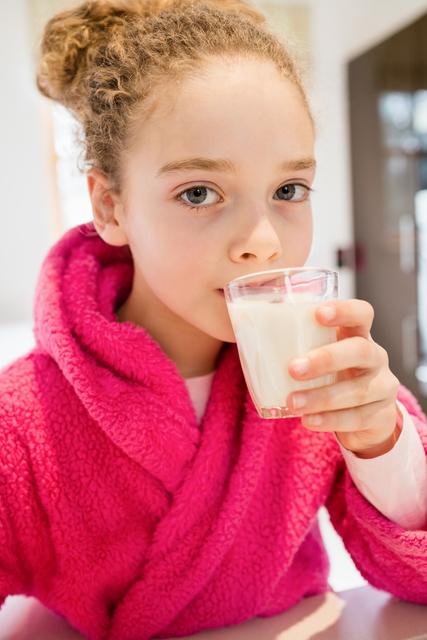 The image shows a young girl in a pink robe drinking a glass of milk in a modern kitchen. This photo can be used for promoting healthy lifestyles, breakfast products, children's nutrition, or for home and family-related content. Ideal for blogs, advertisements, or educational materials focused on healthy eating habits and family routines.