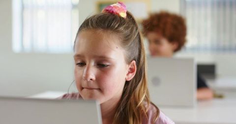 Young girl concentrating on laptop in modern classroom with classmates using laptops