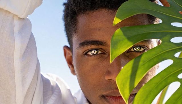 Sunlit portrait of man peeking through monstera leaf with reflective eyes