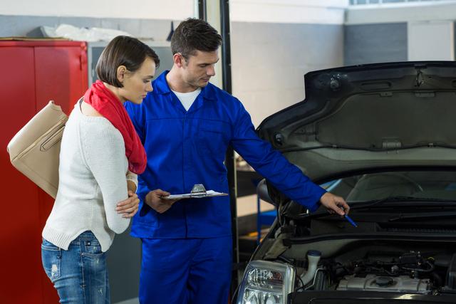 Mechanic in blue uniform explaining car issues to concerned female customer in a repair garage. Ideal for use in content related to automotive services, car maintenance, customer service, and professional mechanics.