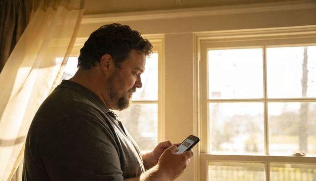 Middle-aged man standing by window using smartphone in warm sunlight