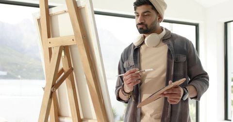 Indian artist painting at easel in bright waterfront studio wearing beanie and headphones