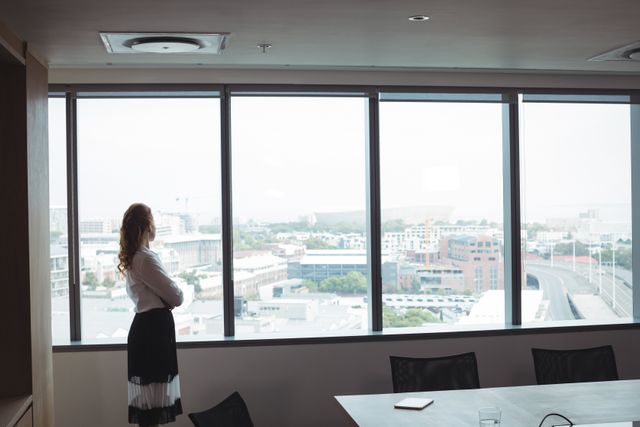Businesswoman standing by office window, looking out at cityscape. Ideal for themes related to business strategy, leadership, corporate environment, professional life, and urban work settings.