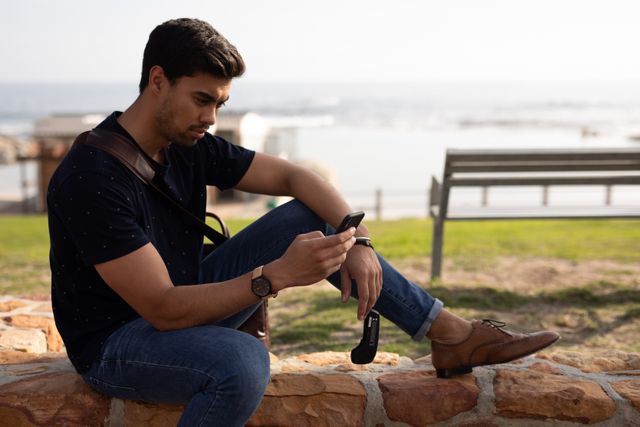 Side view of a biracial man enjoying free time in nature on a sunny day, sitting on the wall with one leg on it, using a smartphone. 