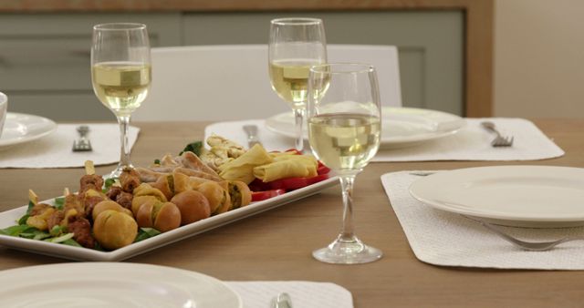 Dinner table set with white plates and glasses of white wine. Appetizers, including skewers and snacks, arranged on a platter. Ideal for illustrating home dining, entertaining, and gourmet food and wine settings. Can be used for blogs, websites, or advertisements related to food, lifestyle, and home gatherings.